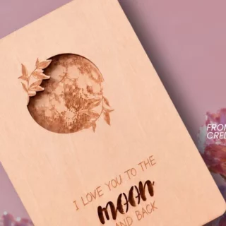 Wooden card with a moon-shaped cutout and floral engraving, reading 'I love you to the moon and back', set against pink flowers.