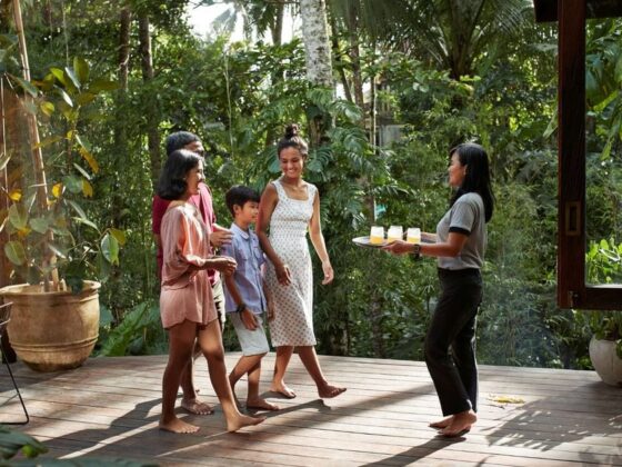 Group of five people on a wooden deck in a lush garden, as a server brings a tray of drinks intact.