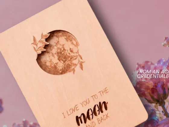 Wooden card with a moon-shaped cutout and floral engraving, reading 'I love you to the moon and back', set against pink flowers.