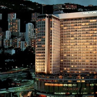 Nighttime cityscape featuring a tall Hilton hotel building illuminated against a backdrop of hills and numerous skyscrapers.