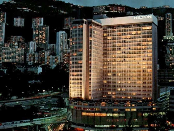 Nighttime cityscape featuring a tall Hilton hotel building illuminated against a backdrop of hills and numerous skyscrapers.