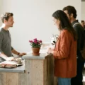 Barista taking an order from a woman at a rustic wooden counter; a potted flower sits on the counter and customers line up behind her in a bright cafe.