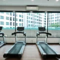 Four treadmills lined up in a bright gym facing large windows with a cityscape view outside. A wall fan and air conditioner are visible.