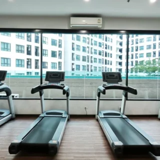 Four treadmills lined up in a bright gym facing large windows with a cityscape view outside. A wall fan and air conditioner are visible.