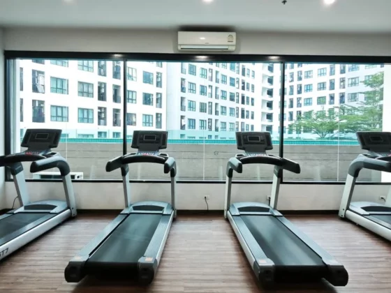 Four treadmills lined up in a bright gym facing large windows with a cityscape view outside. A wall fan and air conditioner are visible.