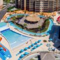Aerial view of a resort waterpark with colorful twists slides, a central thatched hut, and a lazy river surrounded by loungers and buildings.