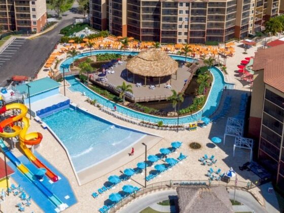 Aerial view of a resort waterpark with colorful twists slides, a central thatched hut, and a lazy river surrounded by loungers and buildings.