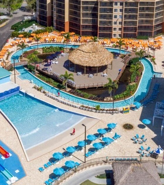 Aerial view of a resort waterpark with colorful twists slides, a central thatched hut, and a lazy river surrounded by loungers and buildings.