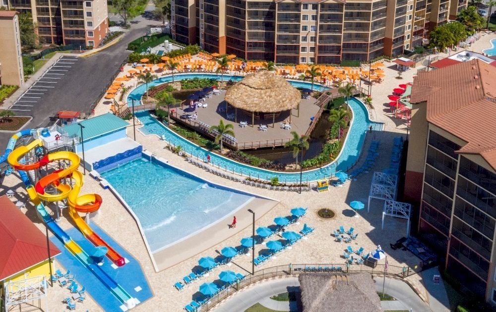 Aerial view of a resort waterpark with colorful twists slides, a central thatched hut, and a lazy river surrounded by loungers and buildings.