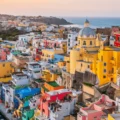 Colorful hillside seaside town with a blue-domed church, pastel houses, and boats in a small harbor at sunset far in the background.