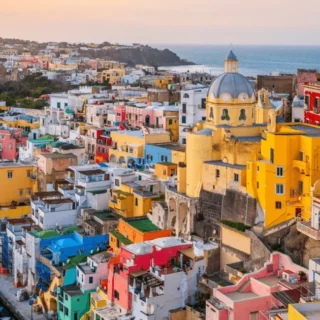 Colorful hillside seaside town with a blue-domed church, pastel houses, and boats in a small harbor at sunset far in the background.