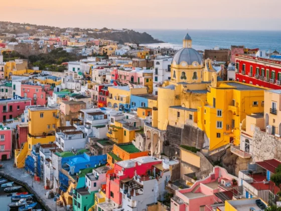 Colorful hillside seaside town with a blue-domed church, pastel houses, and boats in a small harbor at sunset far in the background.