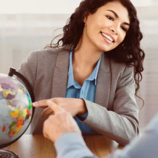 Smiling businesswoman in a gray blazer points to a globe during a meeting with two colleagues at a table