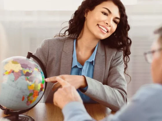 Smiling businesswoman in a gray blazer points to a globe during a meeting with two colleagues at a table