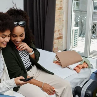 Two friends sit on a bed, smiling while sharing a phone during a travel trip with suitcases nearby.