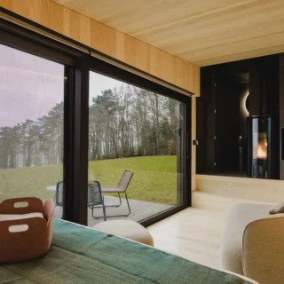 Hotel suite with a floor-to-ceiling glass wall, offering a view of a grassy lawn and trees; bed, seating area, and a fireplace visible inside.