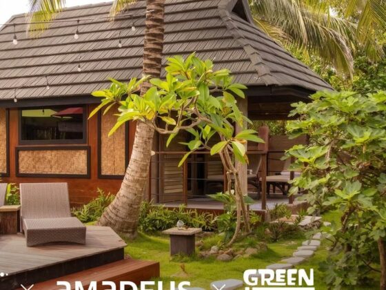 Tropical bungalow with a dark thatched roof, wooden deck, and two wicker lounge chairs amid palm trees and lush greenery.