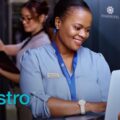 Smiling hotel staffwoman in a blue uniform using a laptop in a server room corridor