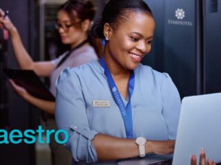Smiling hotel staffwoman in a blue uniform using a laptop in a server room corridor