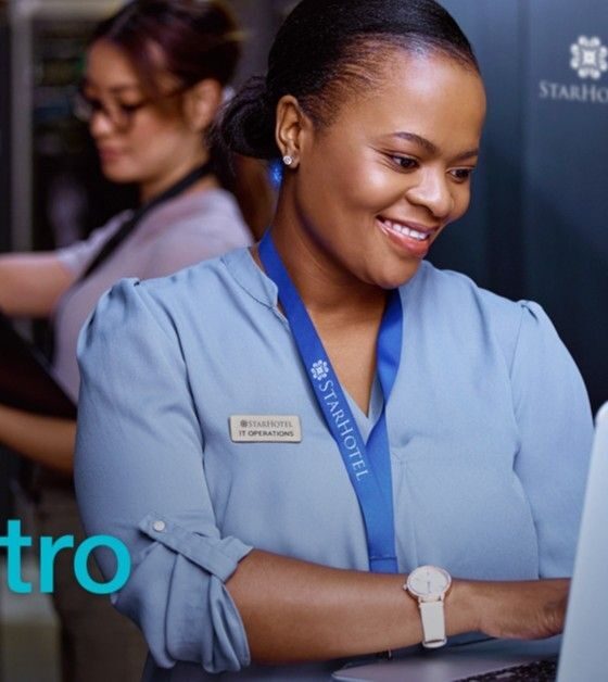 Smiling hotel staffwoman in a blue uniform using a laptop in a server room corridor