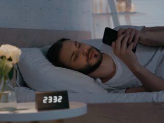 Man lying in bed at night, scrolling on his smartphone; bedside clock shows 5:55 and a white rose in a vase on the nightstand.