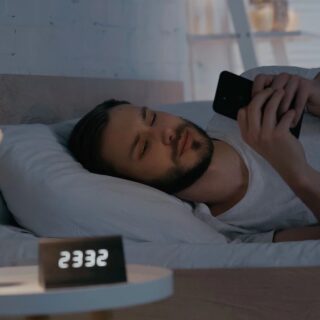 Man lying in bed at night, scrolling on his smartphone; bedside clock shows 5:55 and a white rose in a vase on the nightstand.