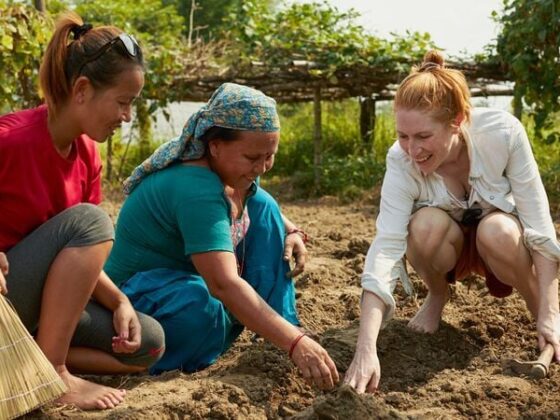 Three women kneeling in a garden, planting seeds and tending rich soil with smiles on a sunny day.