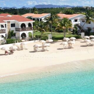 Resort on a white-sand beach with white buildings, red-tiled roofs, palm trees, and rows of umbrellas beside turquoise water