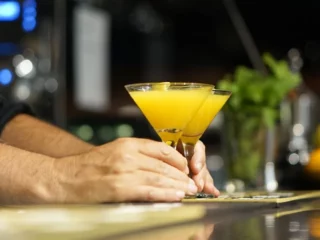 Bartender's hands hold two yellow cocktails in martini glasses over a polished bar counter at a dimly lit bar order scene.