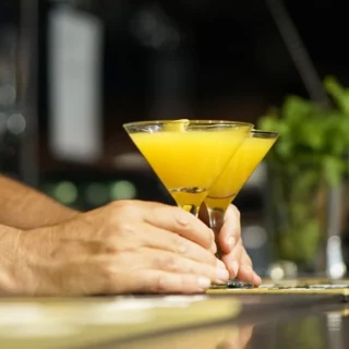 Bartender's hands hold two yellow cocktails in martini glasses over a polished bar counter at a dimly lit bar order scene.