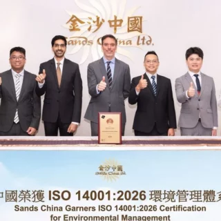 Group of eight professionals posing behind a table, thumbs up, celebrating Sands China Ltd. ISO 14001:2026 certification achievement.