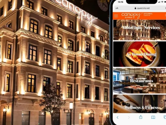 Night view of a warmly lit hotel facade with a smartphone beside it showing the Canopy by Hilton app menu.