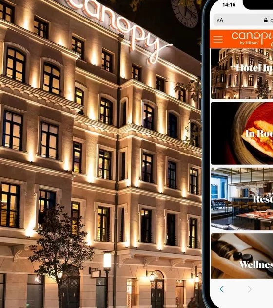 Night view of a warmly lit hotel facade with a smartphone beside it showing the Canopy by Hilton app menu.