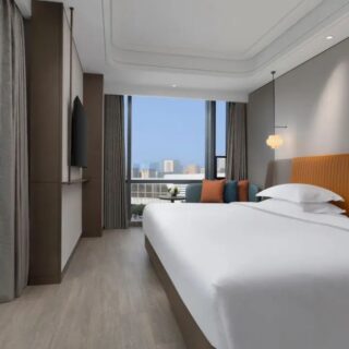 Hotel bedroom with a large white bed and an orange padded headboard, gray walls, and a view of the city skyline through floor-to-ceiling windows.
