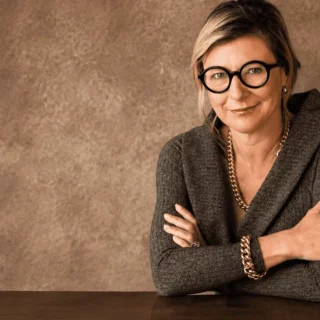Professional woman with short blonde hair and round glasses sits at a wooden table, arms crossed, in a gray blazer.