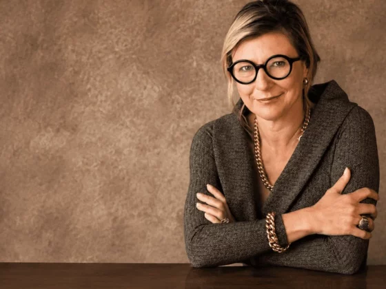 Professional woman with short blonde hair and round glasses sits at a wooden table, arms crossed, in a gray blazer.