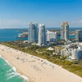 Aerial view of a sunny coastal city with white-sand beach and turquoise water, lined by modern high-rise buildings beyond the shore.