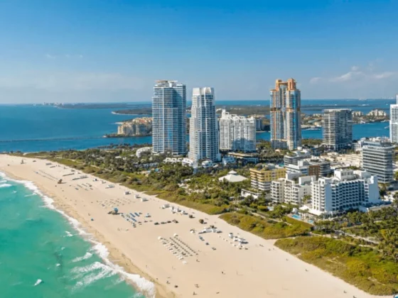 Aerial view of a sunny coastal city with white-sand beach and turquoise water, lined by modern high-rise buildings beyond the shore.
