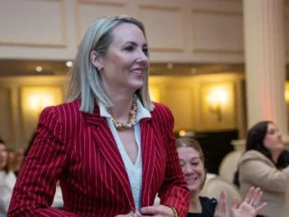 Woman in a red pinstripe blazer smiles as audience applauds at a formal event.