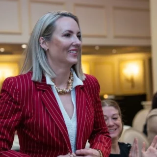 Woman in a red pinstripe blazer smiles as audience applauds at a formal event.