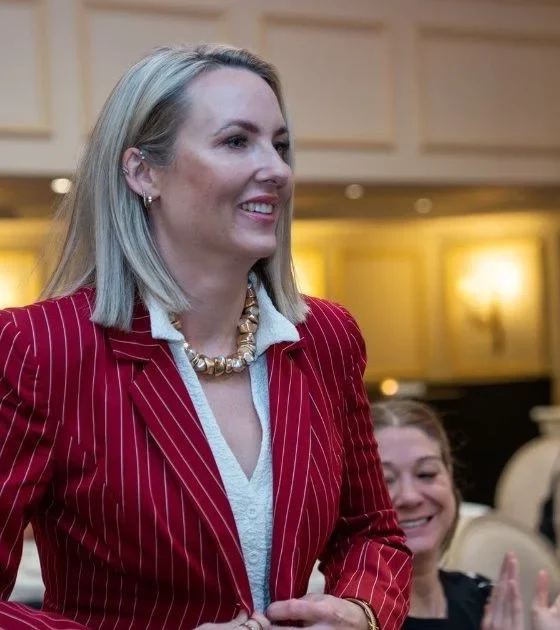 Woman in a red pinstripe blazer smiles as audience applauds at a formal event.