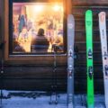 Row of skis propped against a log wall outside a wooden cabin, with snowy ground and warm-lit windows showing people inside.