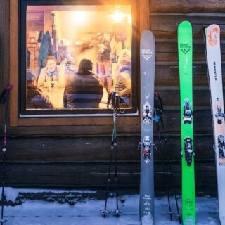 Row of skis propped against a log wall outside a wooden cabin, with snowy ground and warm-lit windows showing people inside.