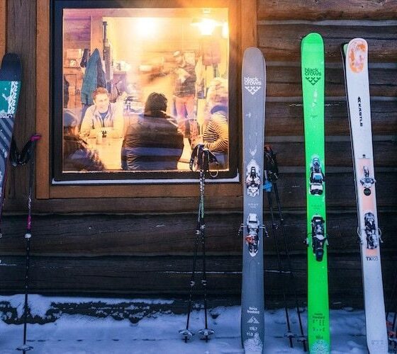 Row of skis propped against a log wall outside a wooden cabin, with snowy ground and warm-lit windows showing people inside.