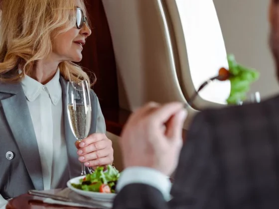 Businesswoman in a gray suit enjoying a glass of champagne and salad on a private jet, wearing sunglasses and looking out the window.
