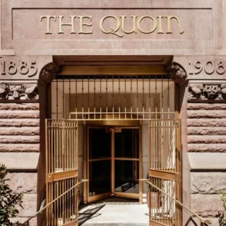 Entrance to The Quoin building with ornate stone facade and open gold gates; the sign reads THE QUOIN above the doorway.