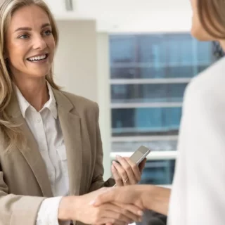 Two professional women in a beige blazer shake hands in a bright office, with one holding a smartphone and smiling.