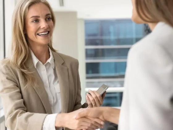 Two professional women in a beige blazer shake hands in a bright office, with one holding a smartphone and smiling.