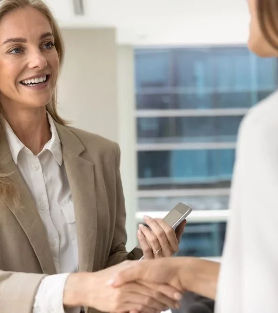 Two professional women in a beige blazer shake hands in a bright office, with one holding a smartphone and smiling.