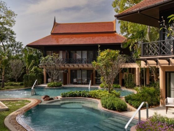 Luxury resort courtyard with a curved pool, tropical plants, and a Thai-style building with a red roof.